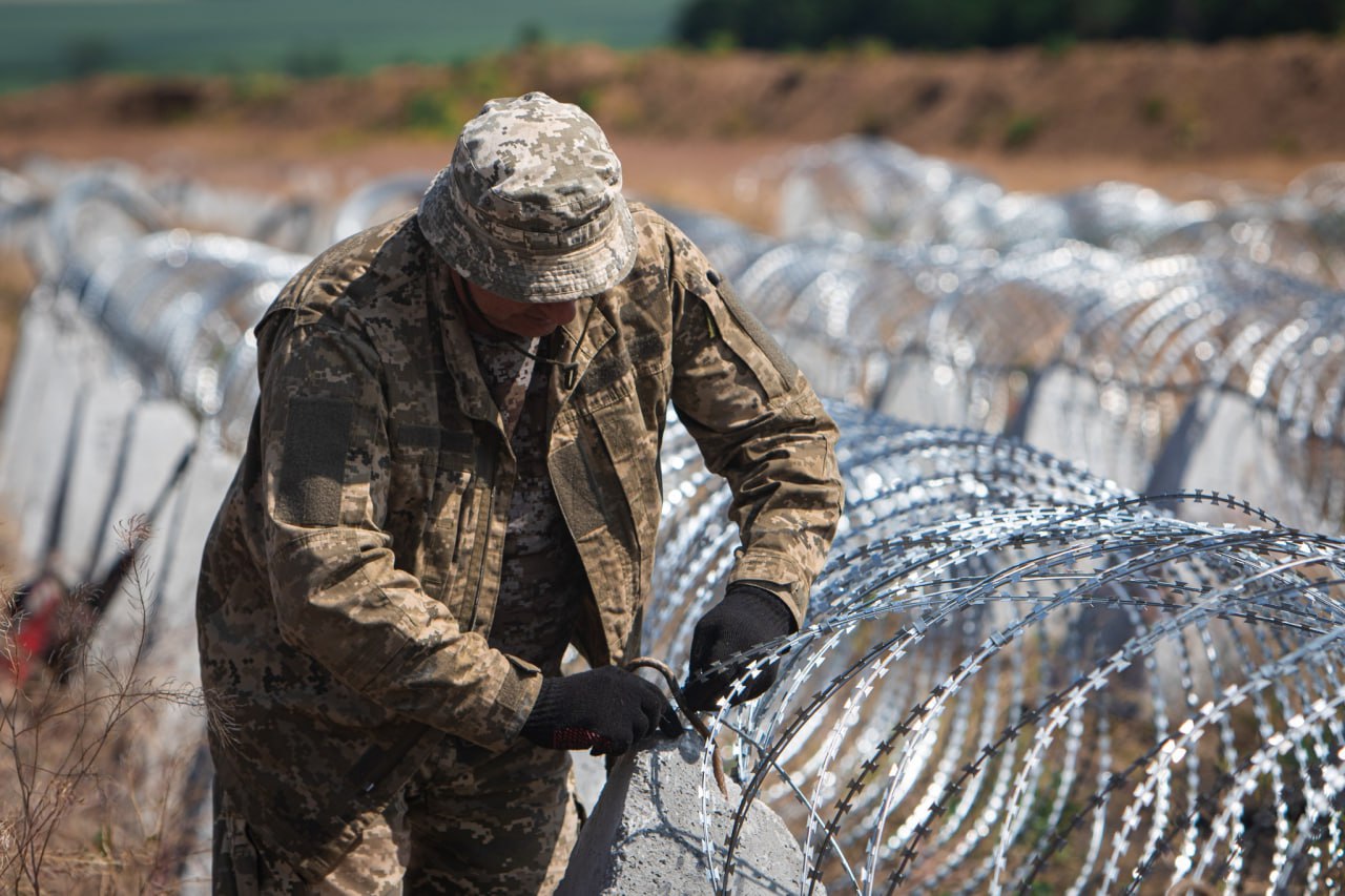 Військовослужбовець у камуфляжній формі та панамі встановлює колючий дріт (спіраль Бруно) на бетонні блоки в полі.