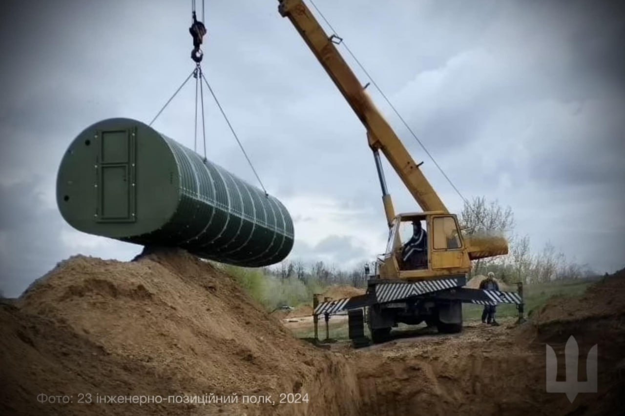 Гусеничний кран опускає велику циліндричну захисну споруду (бункер) зеленого кольору в глибокий котлован.