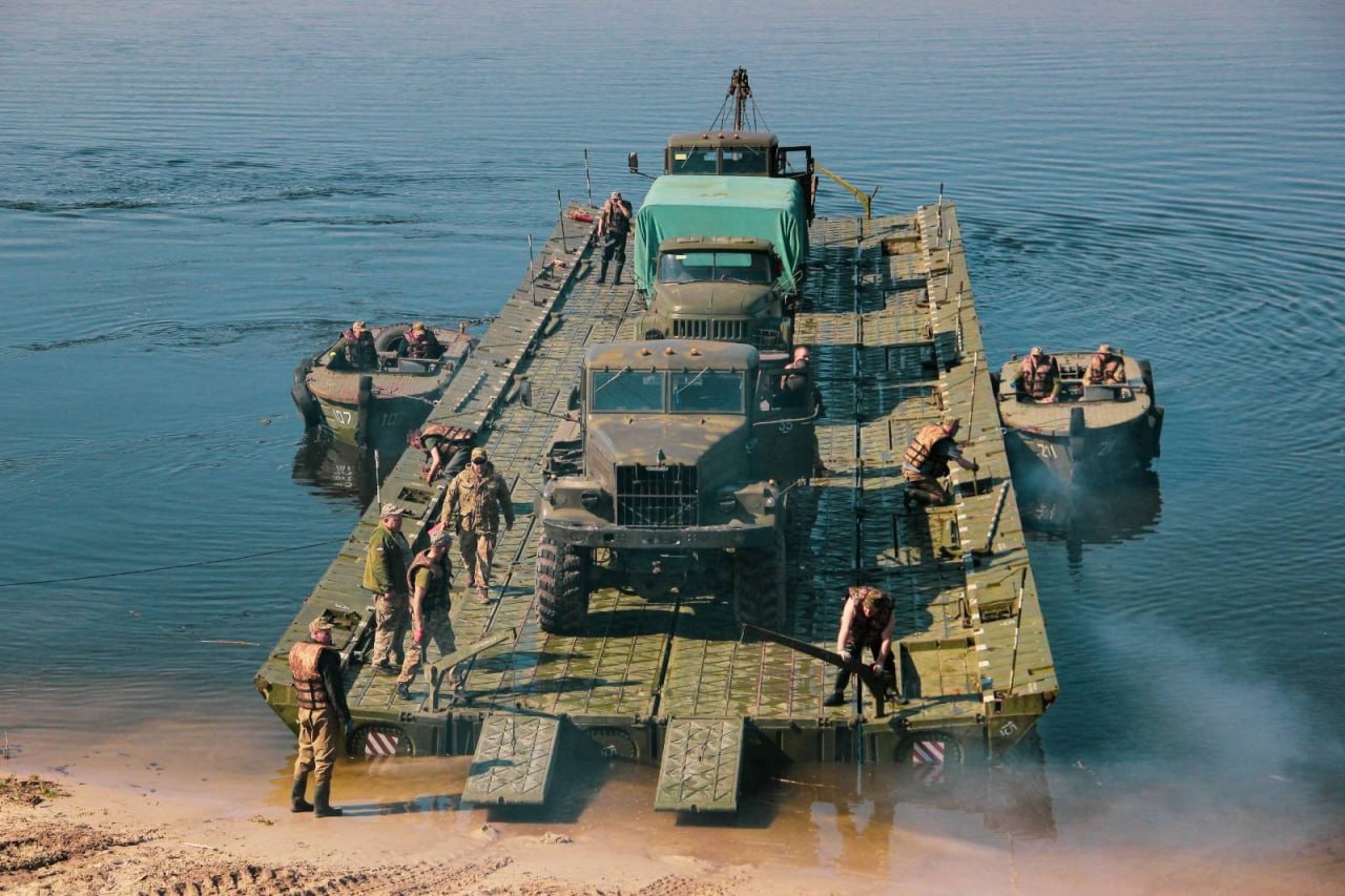 Військовий понтонний пором, зібраний із секцій, перевозить дві вантажівки та військовослужбовців через річку, швартуючись до берега.
