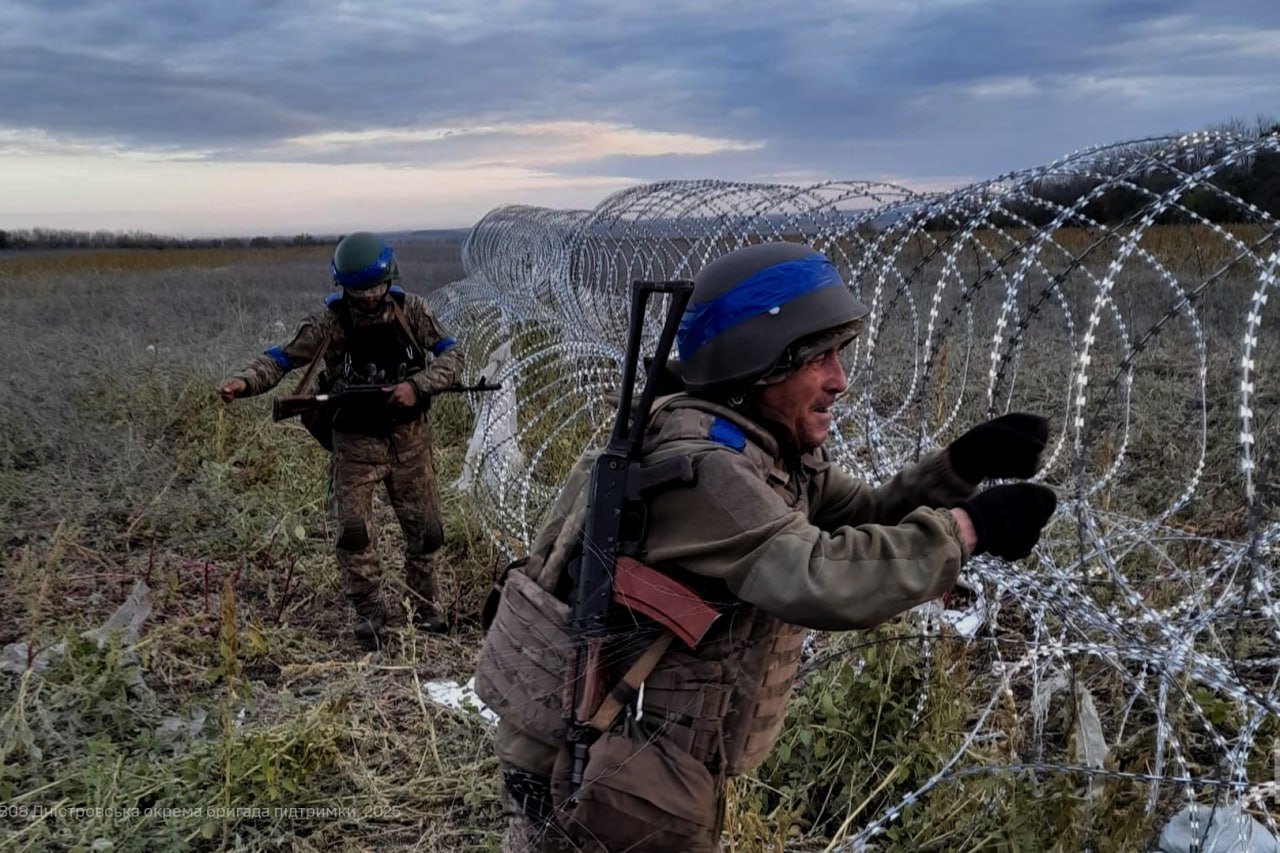 Два українські військовослужбовці в польовій формі та шоломах, обмотаних синьою стрічкою, встановлюють або долають спіральне загородження з колючого дроту на відкритій місцевості під похмурим небом.
