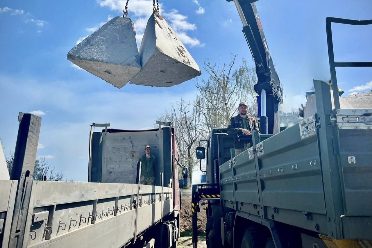 Два українські військовослужбовці стоять на вантажних автомобілях, спостерігаючи, як кран-маніпулятор піднімає в повітря дві великі бетонні протитанкові піраміди, відомі як