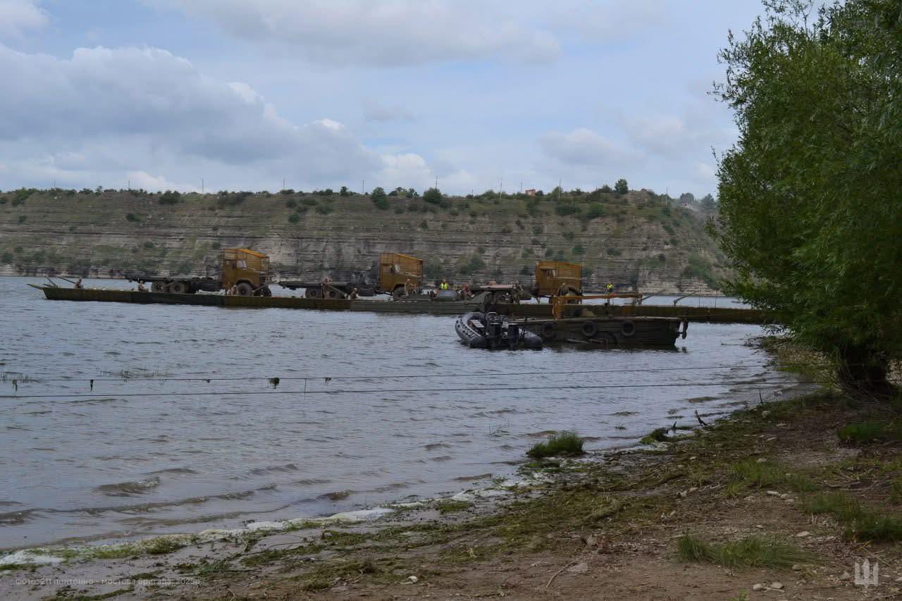 На водоймі видно понтонний міст, що складається з кількох секцій, на яких розміщені великі вантажні автомобілі (ймовірно, військові). Зображення зроблене з берега, на передньому плані видно частину берегової лінії з деревом, а на задньому плані — високий пагорб з жовто-сірими скелями під хмарним небом. Ймовірно, це навчання або розгортання понтонно-мостового підрозділу.