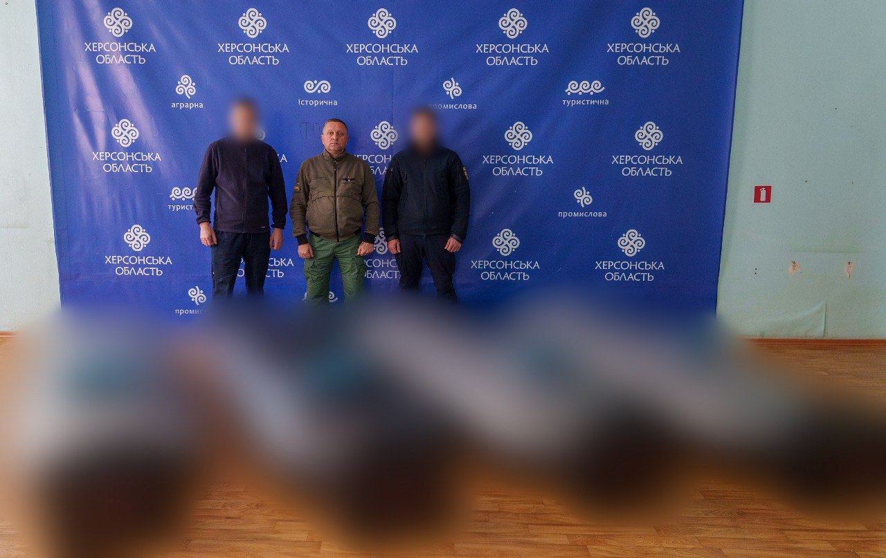Троє чоловіків, обличчя яких частково заблюрені, стоять перед синім прес-волом з логотипами та текстом ХЕРСОНСЬКА ОБЛАСТЬ. Центральний чоловік одягнений у захисну куртку, двоє інших – у темний одяг.