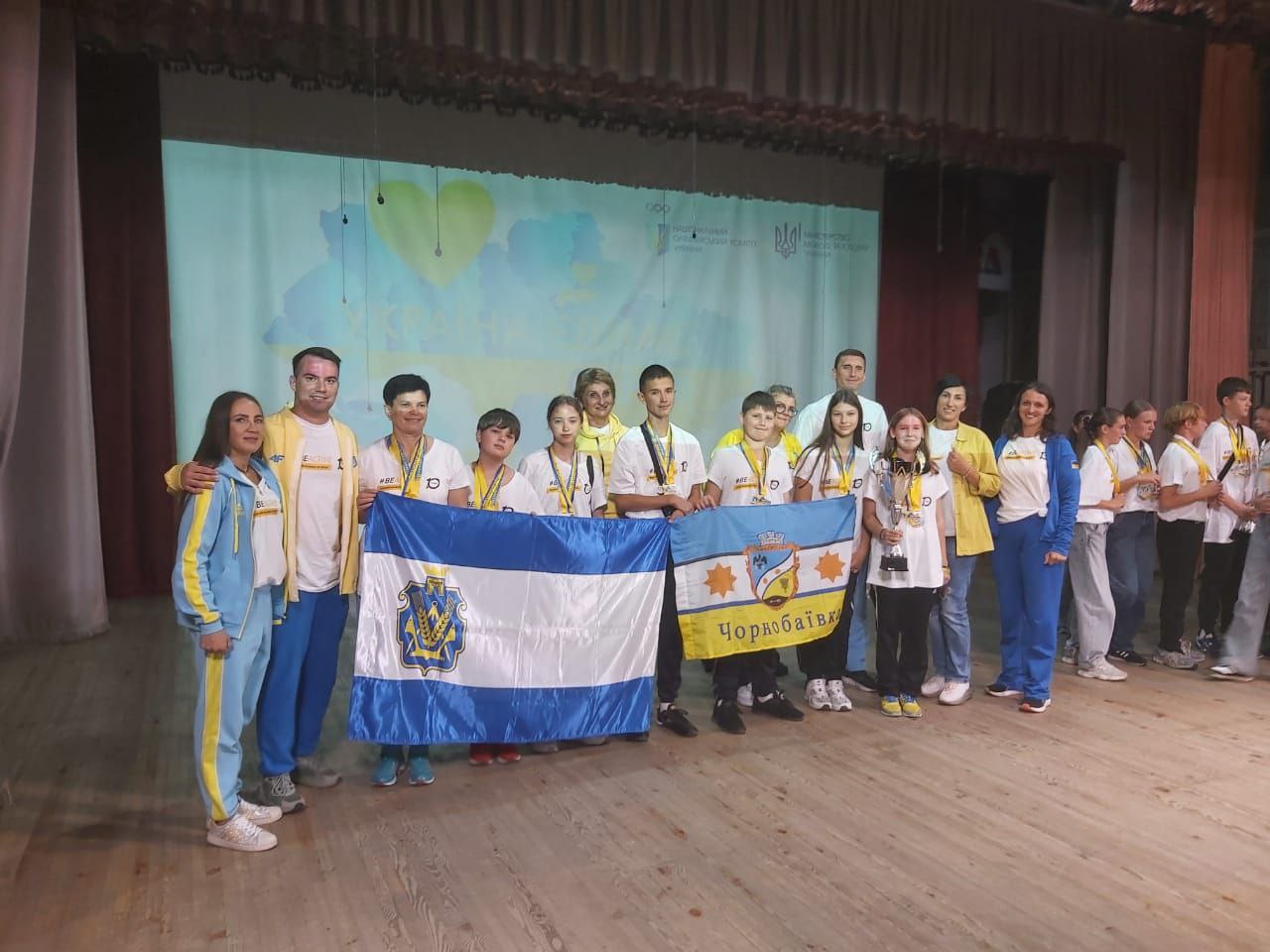 Група молоді та дорослих стоїть на сцені в спортивному одязі (переважно біло-жовто-синіх кольорів) із медалями та кубками. Вони тримають синьо-білий прапор із гербом та інший прапор з написом Чорнобаївка на тлі проекції із зображенням серця та символікою України.