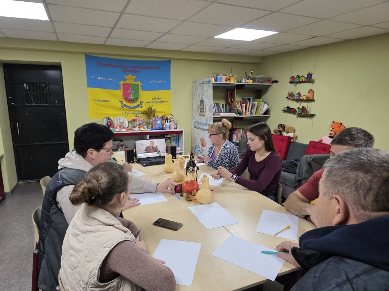 Група з шести людей сидять за столом у приміщенні та беруть участь в обговоренні чи навчанні. На столі лежать чисті аркуші паперу та кілька гарбузів, а на задньому плані видно прапор Пересадівської територіальної громади та ноутбук з відеодзвінком.