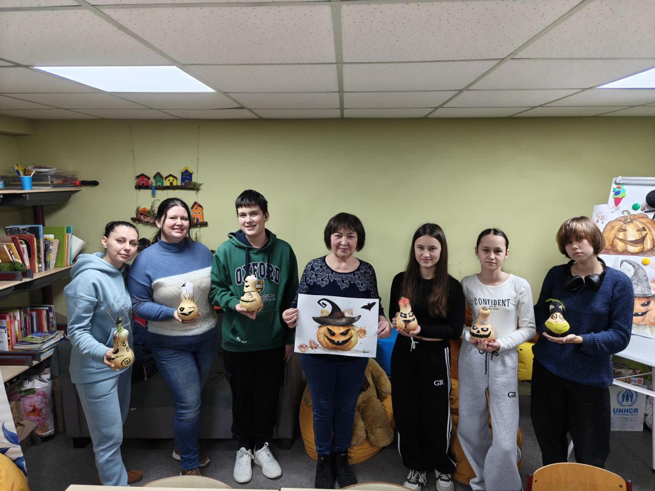 Сім людей стоять у приміщенні та показують свої вироби ручної роботи до Хелловіну: розписані маленькі фігурки гарбузів і плакат із зображенням гарбуза-ліхтаря.