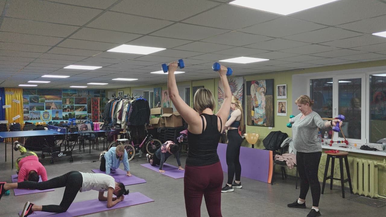 Група з шести жінок займаються фітнесом у приміщенні. Вони виконують різні вправи на фіолетових килимках, деякі використовують легкі гантелі, а одна жінка робить вправу відведення ноги на килимку. На задньому плані видно столи для настільного тенісу та різноманітні речі.