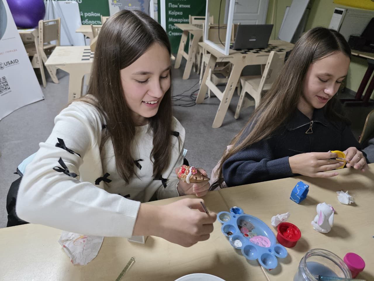 Дві молоді дівчини (підлітки) сидять за столом і посміхаються, займаючись творчістю. Вони розмальовують маленькі керамічні фігурки, використовуючи пензлі та палітру з фарбами.