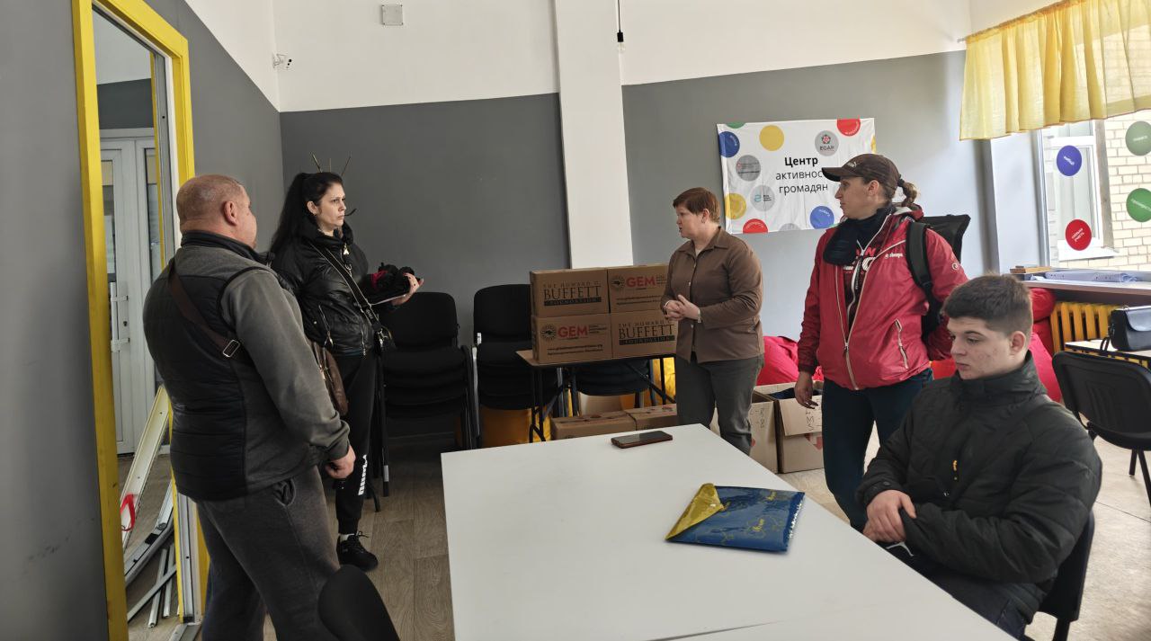 П'ятеро людей знаходяться в приміщенні, яке схоже на Центр активних громадян, про що свідчить плакат на стіні. Дві жінки розмовляють біля столу, на якому стоять картонні коробки з написом RUFFETT, а двоє чоловіків і ще одна жінка стоять або сидять поруч. На столі лежить маленький український прапор.