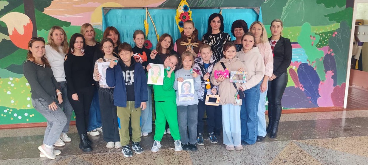 Група з приблизно 16 дорослих жінок і дітей стоїть разом для групового фото на тлі яскравого мальовничого панно. Кілька дітей у центрі групи тримають свої малюнки або вироби ручної роботи.