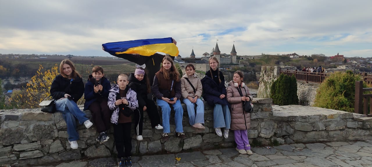 Група з восьми дітей та підлітків сидять або стоять на кам'яній стіні, дивлячись у камеру. На задньому плані видно Кам'янець-Подільську фортецю, а над ними розгорнуто великий прапор України.