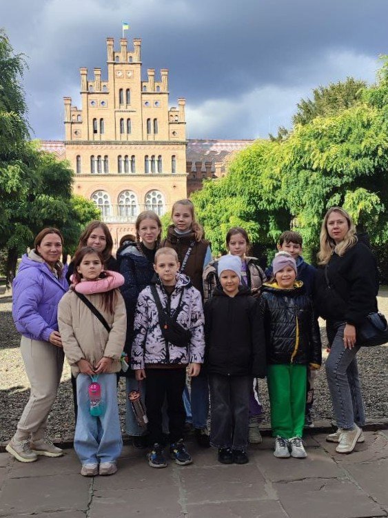 Група з десяти дітей та двох дорослих жінок стоять для групового фото на відкритому повітрі. На задньому плані видно величну старовинну будівлю в неоготичному стилі, оточену деревами (ймовірно, Чернівецький національний університет).