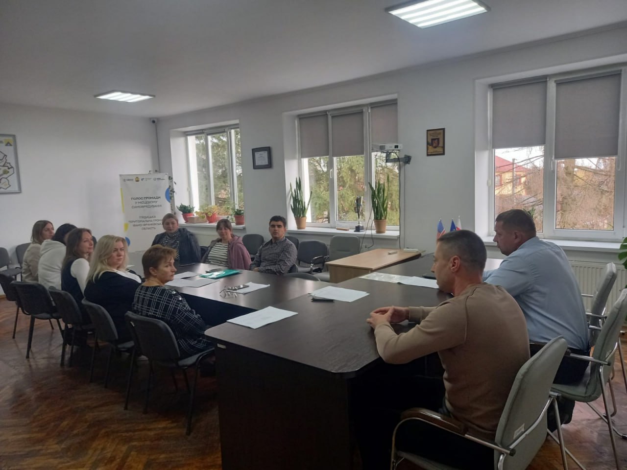 Група з десяти людей (вісім жінок і двоє чоловіків) сидять за великим столом для нарад у світлому офісному приміщенні з великими вікнами та обговорюють документи, що лежать на столі. На задньому плані видно банер з написом: ГОЛОС ГРОМАДИ В ОРГАНАХ САМОВРЯДУВАННЯ.