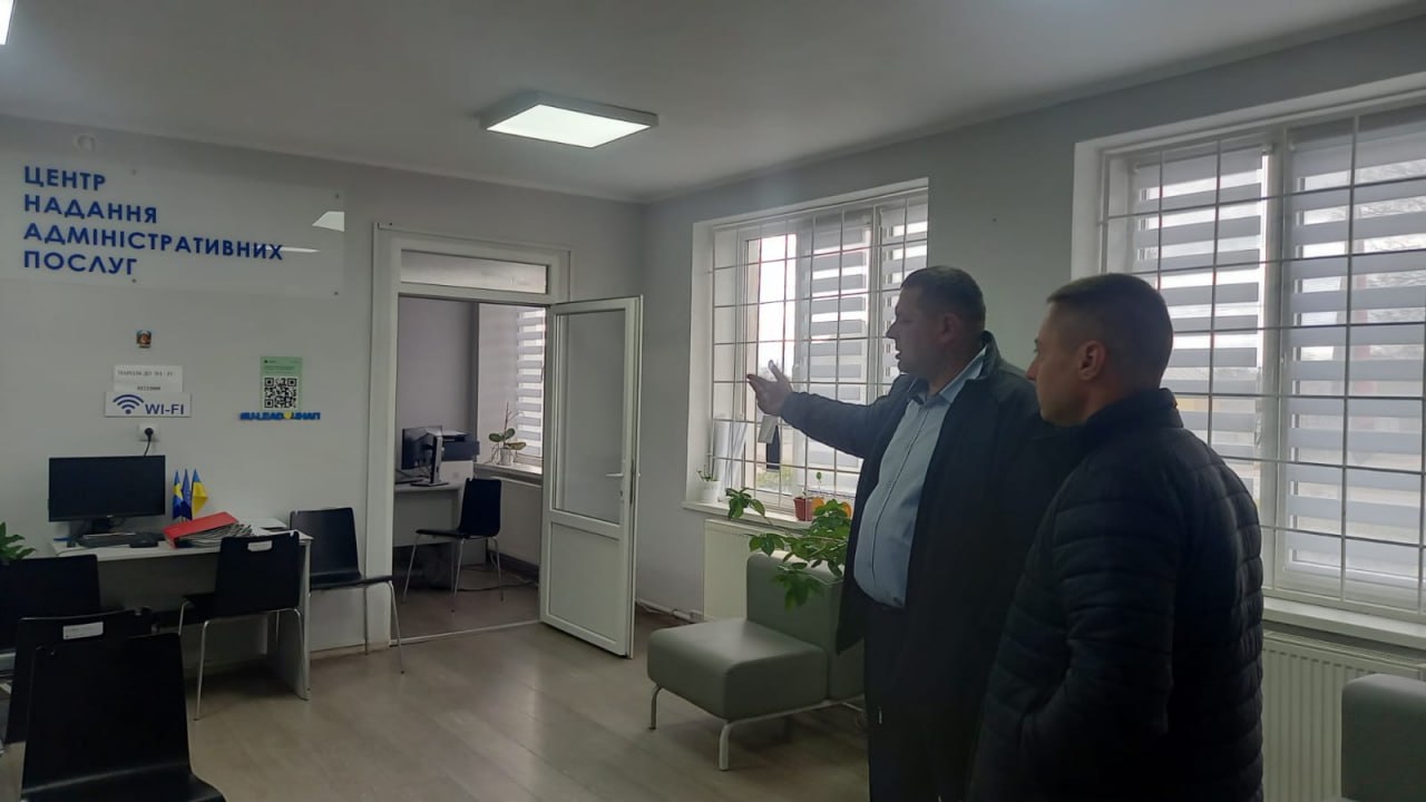 Двоє чоловіків стоять у приміщенні, схожому на Центр надання адміністративних послуг (ЦНАП), про що свідчить напис на стіні. Один чоловік вказує рукою на щось, розмовляючи з іншим чоловіком на тлі робочих місць та великих вікон.