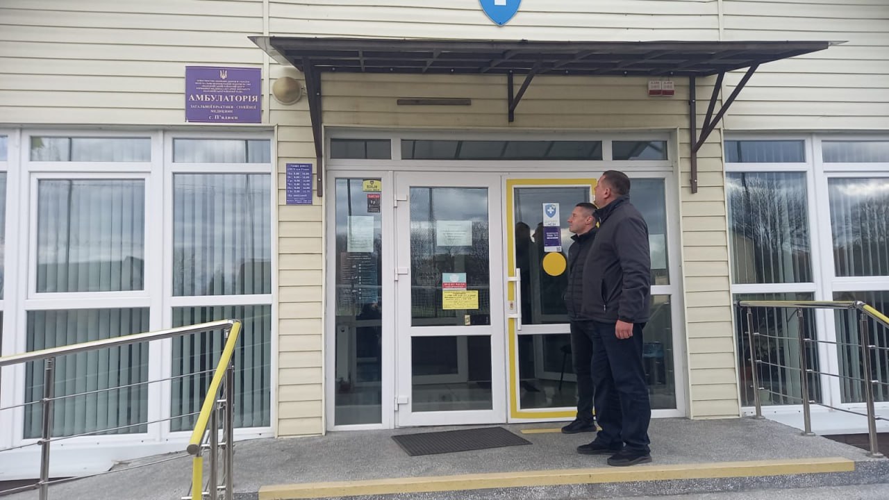 Двоє чоловіків стоять біля входу до будівлі Амбулаторії загальної практики – сімейної медицини в населеному пункті. Будівля має великі вікна та обладнана пандусом.