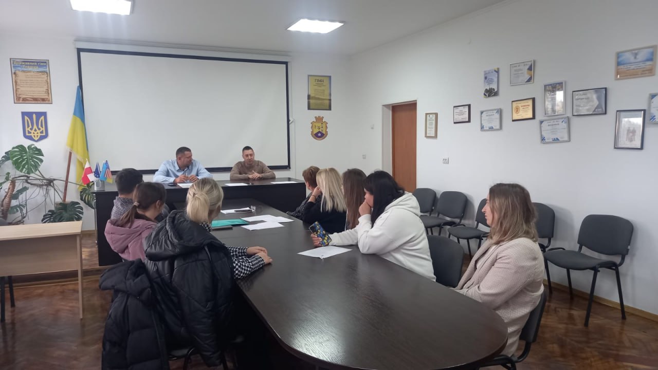 Група людей, переважно жінок, сидять за довгим столом у конференц-залі та беруть участь у нараді чи презентації. Двоє чоловіків сидять на чолі столу перед великим білим проєкційним екраном. У кімнаті також видно український прапор та численні сертифікати/подяки на стінах.