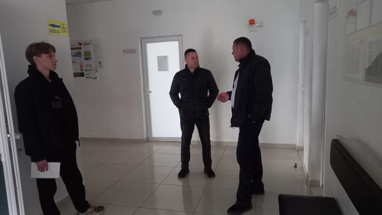 Троє людей (одна жінка та двоє чоловіків) стоять та спілкуються в світлому коридорі офісної будівлі або адміністративної установи. На стіні за ними є інформаційні стенди та вказівник.