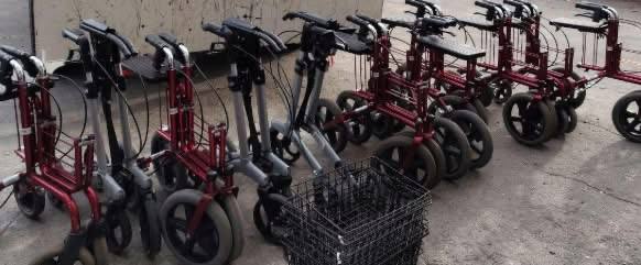 Ряд червоних і сірих ходунків-роляторів (реабілітаційних візків) з сидіннями та кошиками, вишикувані на сірому асфальті.