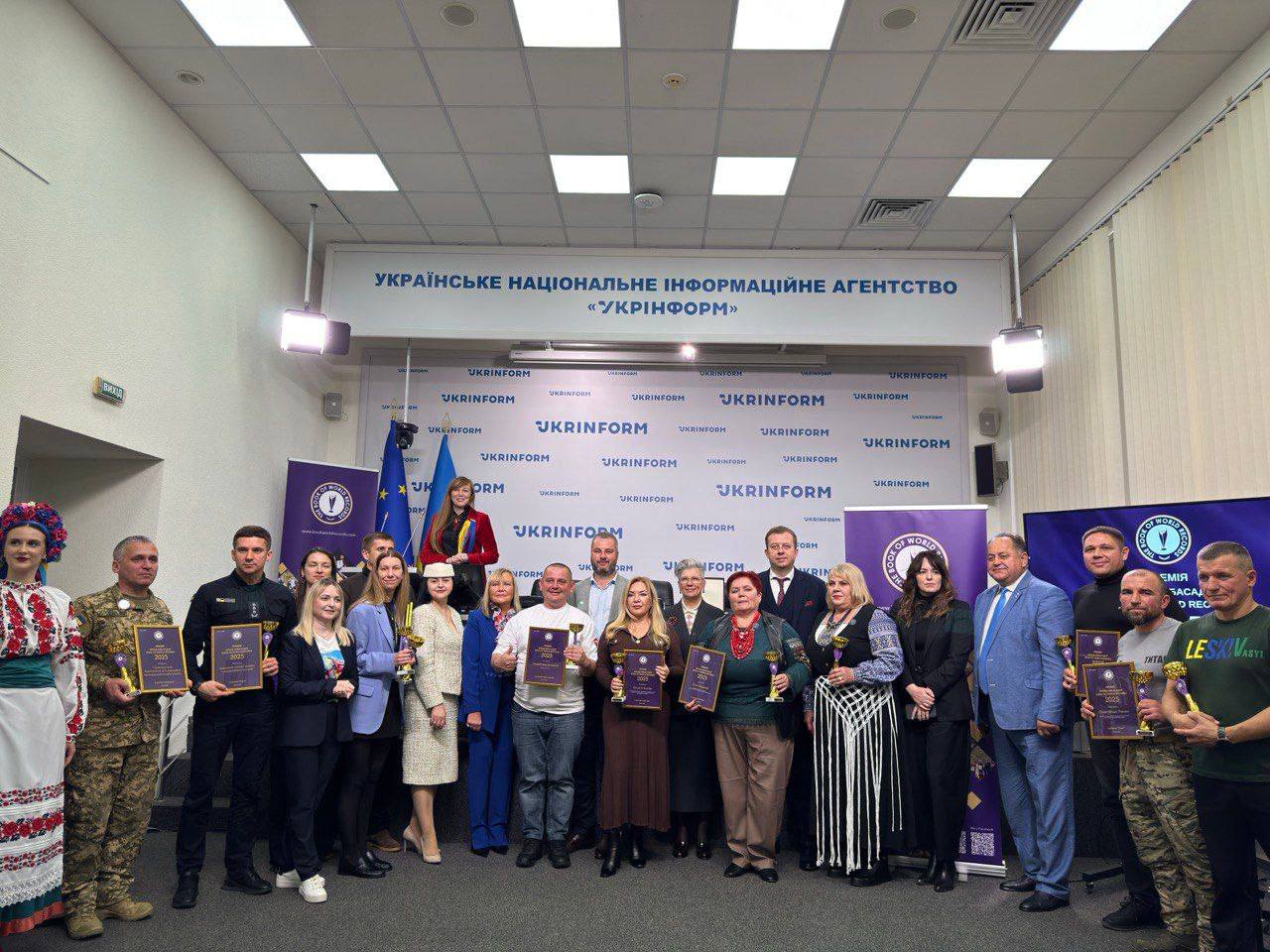 Групове фото близько 25 людей, які стоять перед банером: Українське національне інформаційне агентство УКРІНФОРМ. Вони одягнені у різний одяг (від військової форми та вишиванки до ділових костюмів) та тримають у руках нагороди, кубки та сертифікати після церемонії нагородження.