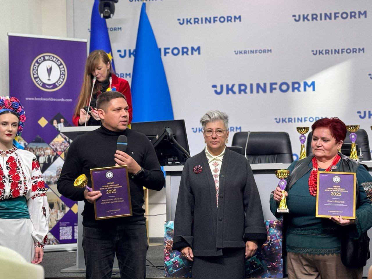 Четверо людей стоять на сцені або біля банера: УКРІНФОРМ після церемонії нагородження. Чоловік у центрі з мікрофоном, жінка ліворуч у вишиванці, а також жінка праворуч тримають нагороди (кубки та дипломи з написом Героїка областей України 2025).