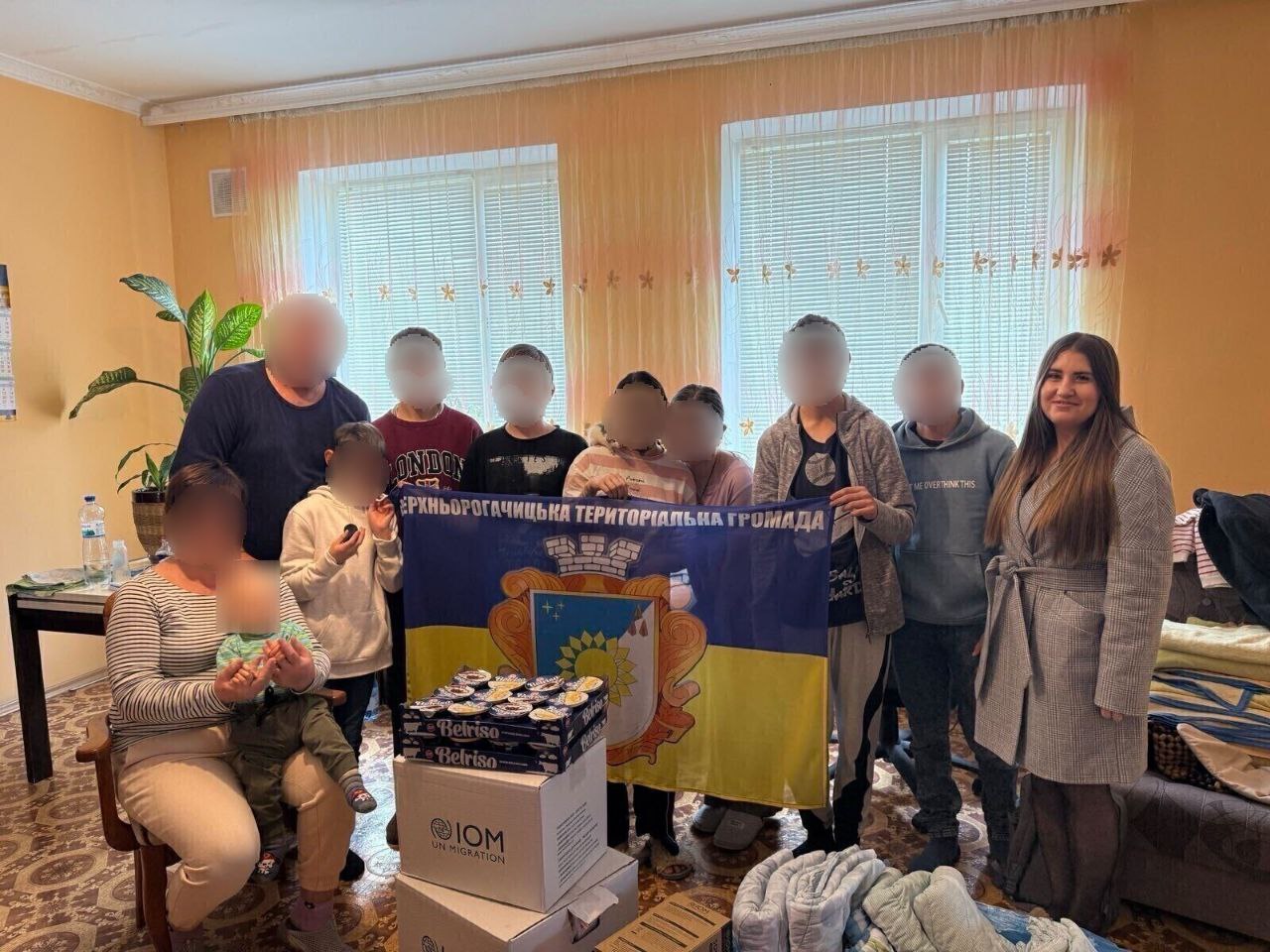 Група з приблизно дев'яти людей, включаючи дорослих, підлітків та маленьку дитину, стоять у житловому приміщенні та тримають великий синьо-жовтий прапор Верхньорогачицької територіальної громади. Перед ними стоять коробки з гуманітарною допомогою, одна з яких містить логотип IOM (Міжнародна організація з міграції).