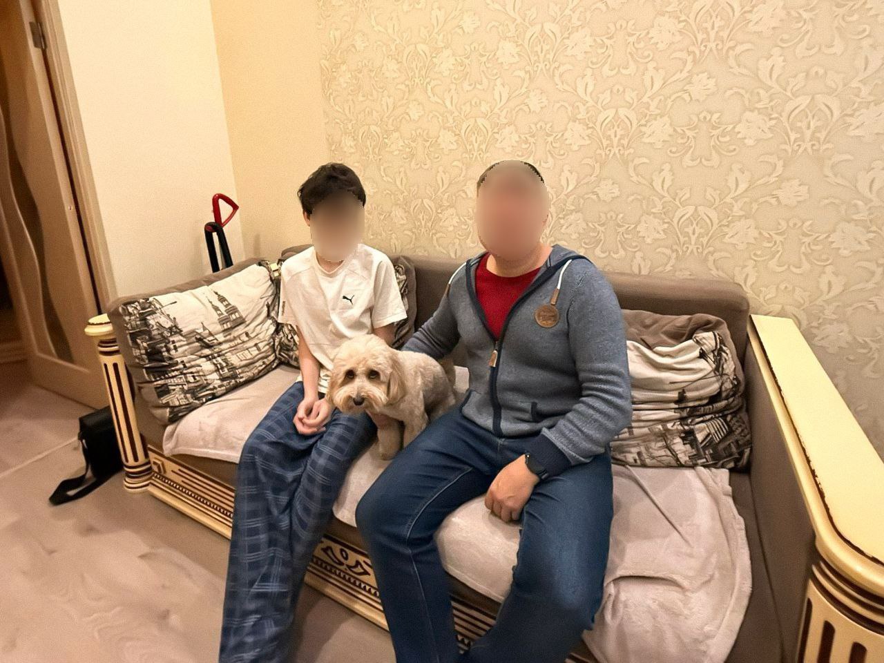 Чоловік і хлопчик (з розмитими обличчями) сидять разом на дивані в кімнаті, а між ними сидить маленька світла кудлата собака.