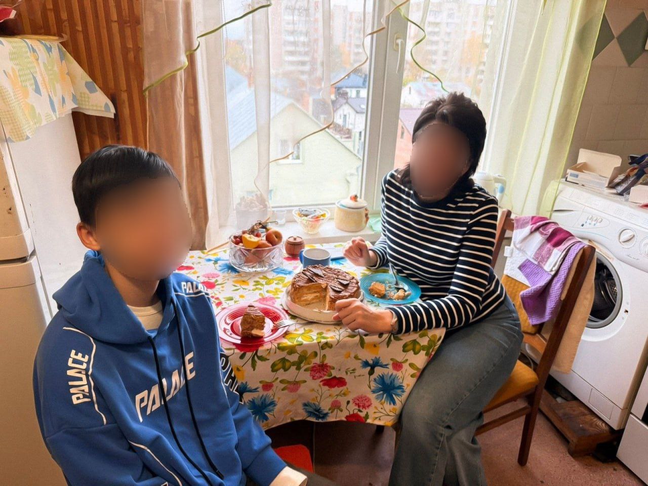 Хлопчик у синьому худі та жінка в смугастій кофті сидять за кухонним столом біля вікна, їдять торт. На столі також є тарілка з фруктами.