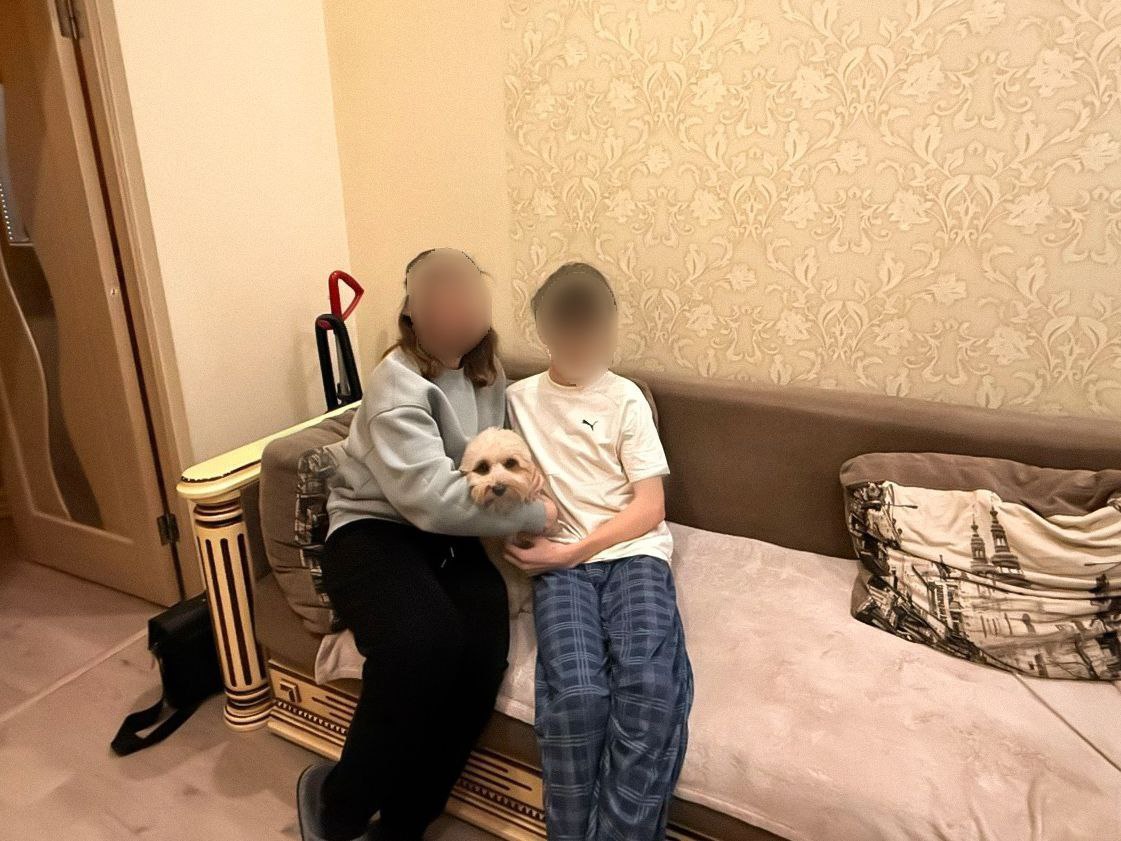 Жінка та хлопчик сидять поруч на дивані у вітальні, тримаючи на руках маленьку світлу собаку. На задньому плані видно світлі шпалери з візерунком.