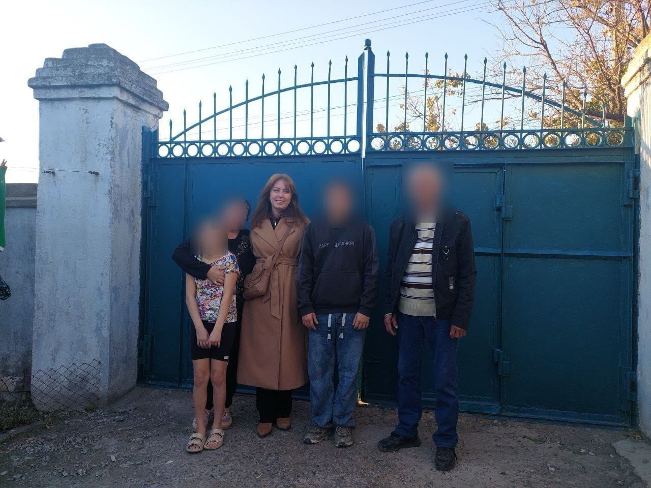 Група з чотирьох людей, включаючи жінку у світло-коричневому пальті та двох чоловіків і дівчинку, стоїть перед високими блакитними металевими воротами на вулиці у сонячний день.