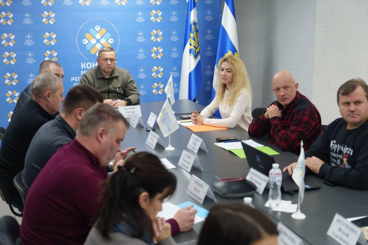 Груповий знімок з офіційної зустрічі або наради, де близько десяти осіб, включаючи чоловіків у військовій та цивільній формі, а також жінок, сидять за довгим столом. На столі розташовані прапорці, ноутбуки та папери. Фон прикрашений синіми банерами з емблемою та державними прапорами, що вказує на офіційний захід.