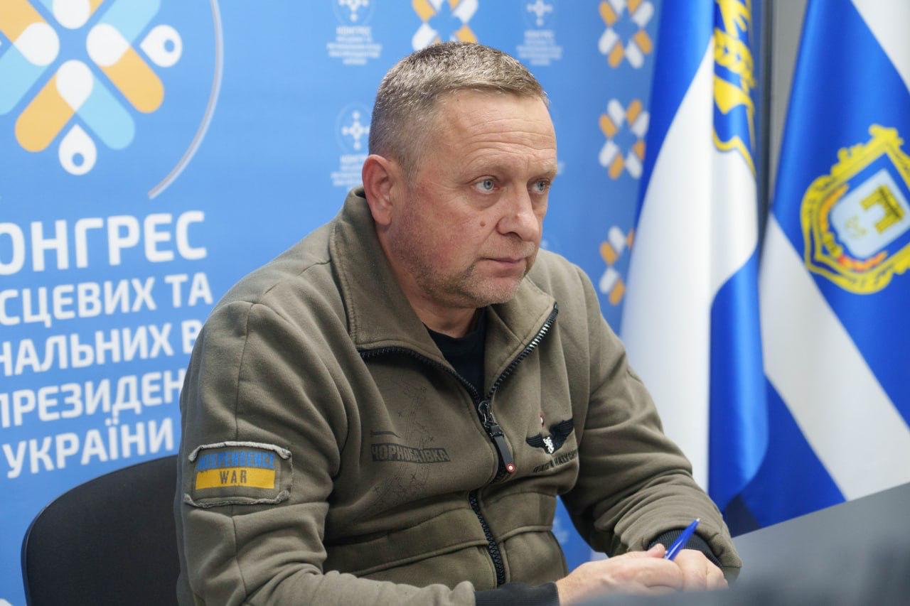 Портретний знімок чоловіка середнього віку у військовій куртці-флісці, що сидить за столом під час наради. На його куртці видно нашивку WAR. На задньому плані – синій банер з написом: КОНГРЕС МІСЦЕВИХ ТА РЕГІОНАЛЬНИХ РАД ПРЕЗИДЕНТА УКРАЇНИ та прапори (зокрема, прапор України).