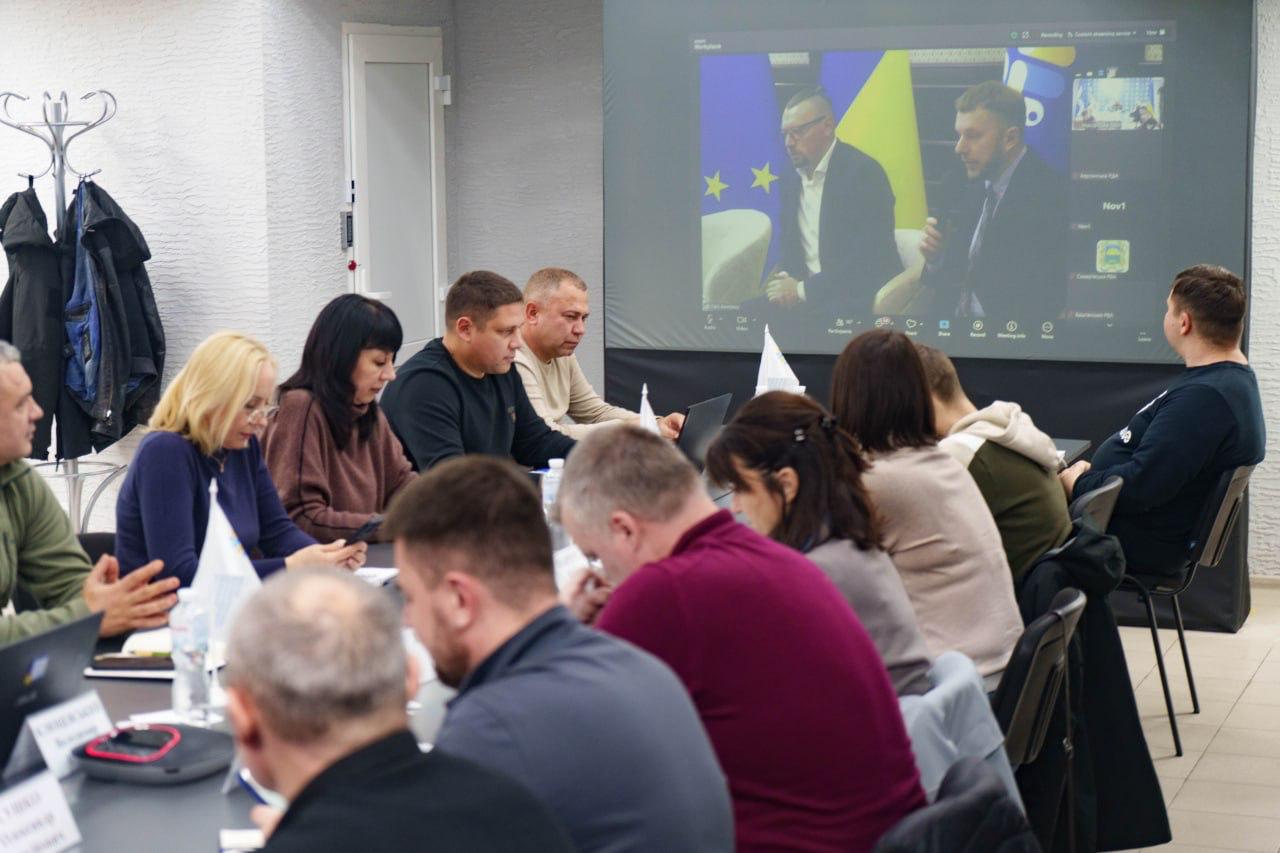 Груповий знімок з наради, де близько десяти осіб, сидячи за столом, дивляться на великий настінний екран. На екрані відбувається відеоконференція за участю двох чоловіків на тлі прапорів України та Європейського Союзу. Учасники наради одягнені у цивільний та військовий одяг, обговорюючи питання за столом.