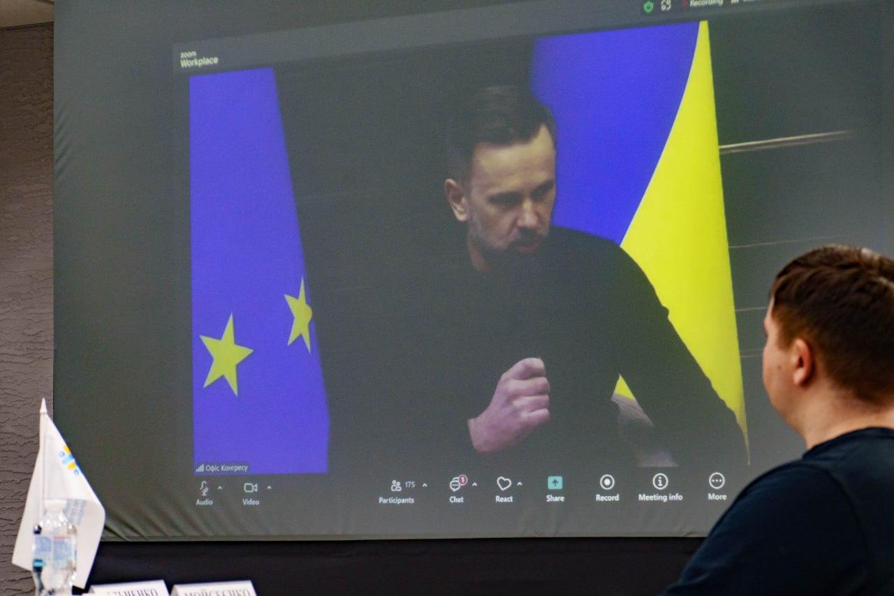 Знімок екрана, на якому відображається відеоконференція з чоловіком у темному светрі, який жестикулює, сидячи на тлі прапорів Європейського Союзу (ліворуч) та України (праворуч). У нижній частині екрана видно елементи інтерфейсу відеодзвінка.