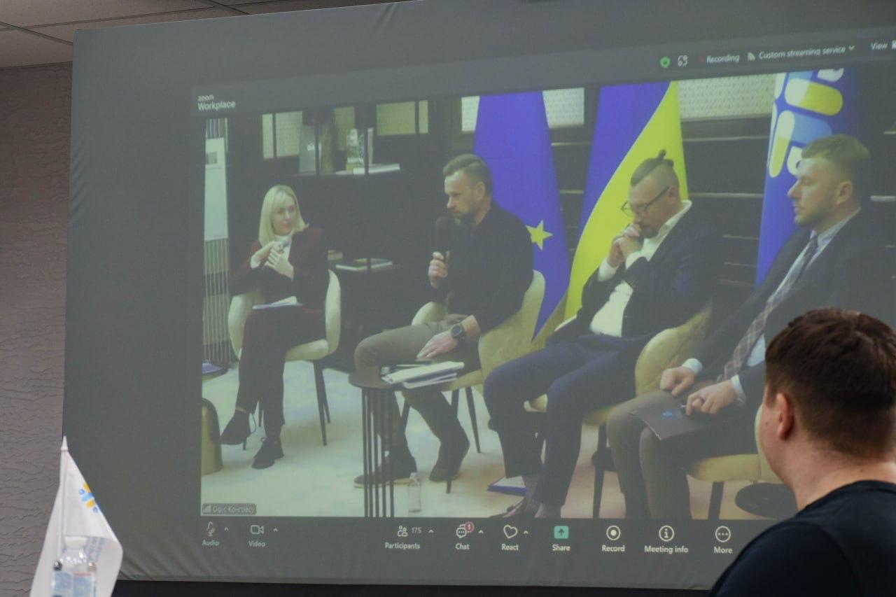 Знімок екрана, на якому транслюється відеоконференція з чотирма учасниками (одна жінка та троє чоловіків) у студії. Учасники сидять на стільцях, двоє з чоловіків тримають мікрофони. Фон студії прикрашений прапорами України та Європейського Союзу. На передньому плані видно потилицю людини, яка спостерігає за трансляцією.