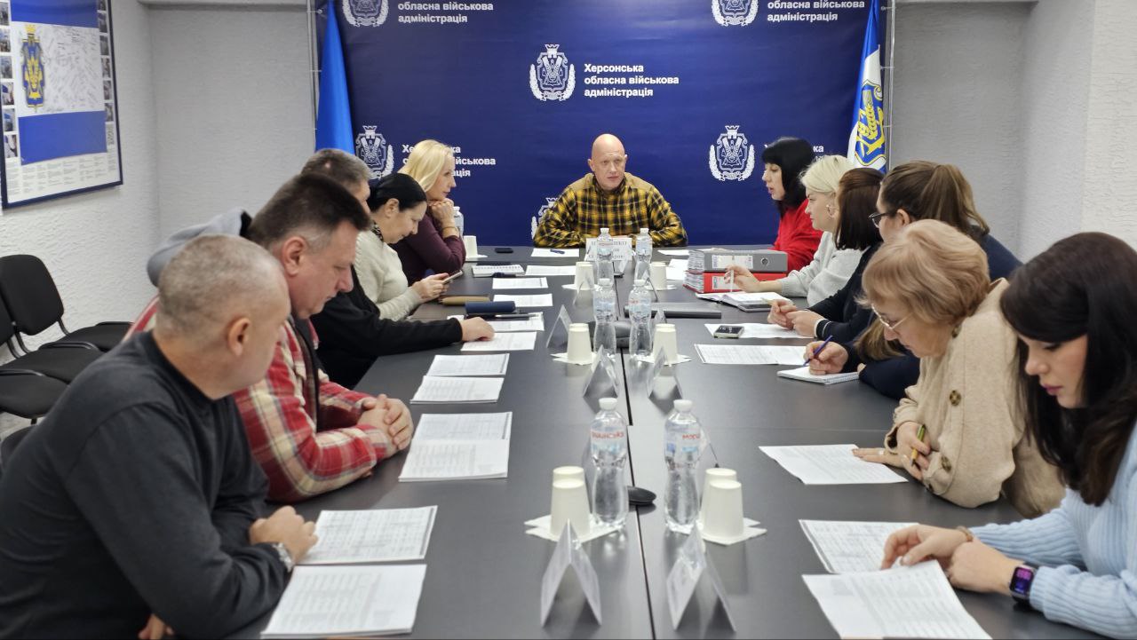 Група людей сидить за довгим столом у конференц-залі Херсонської обласної військової адміністрації (ХОВА), на тлі банера з логотипом адміністрації. У центрі сидить лисий чоловік у картатій сорочці, звертаючись до присутніх.
