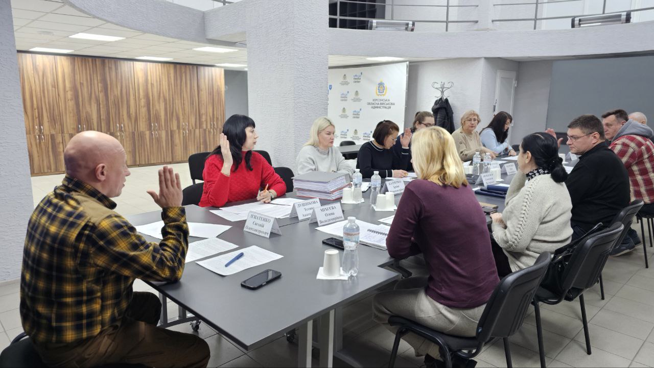 Група людей сидить навколо прямокутного столу в офісному приміщенні. Чоловік у картатій сорочці зліва підняв праву руку, звертаючись до жінки в червоному светрі. На столі видно таблички з іменами та документи.