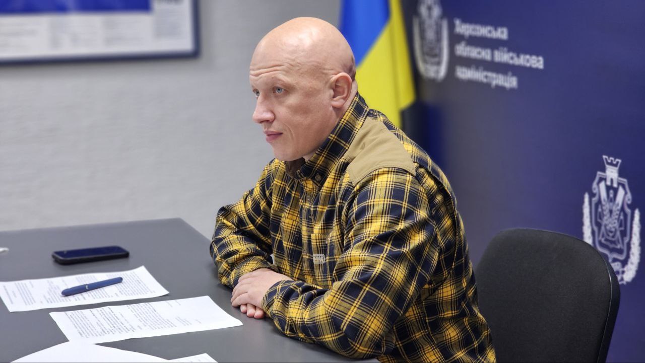Крупний план лисого чоловіка у жовто-чорній картатій сорочці, який сидить за столом під час зустрічі. На задньому плані видно частину банера з написом «Херсонська обласна військова адміністрація» та прапор України.