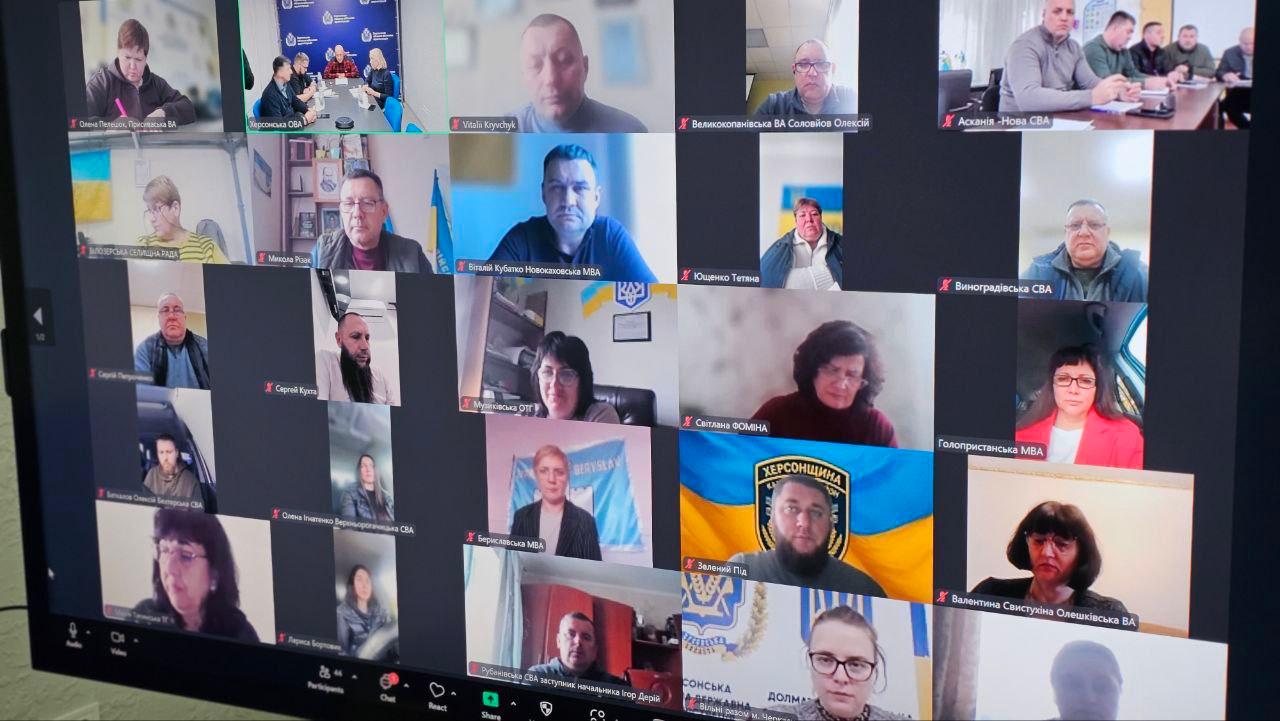 Знімок екрана онлайн-конференції (відеозустрічі) з великою кількістю учасників, розташованих у сітці. На одному з вікон видно людей, які сидять за столом (схоже на учасників з попереднього зображення). Деякі вікна містять логотипи або написи, як-от Херсонщина, СВА (сільська військова адміністрація), МВА (міська військова адміністрація) та Херсонська обласна військова адміністрація.