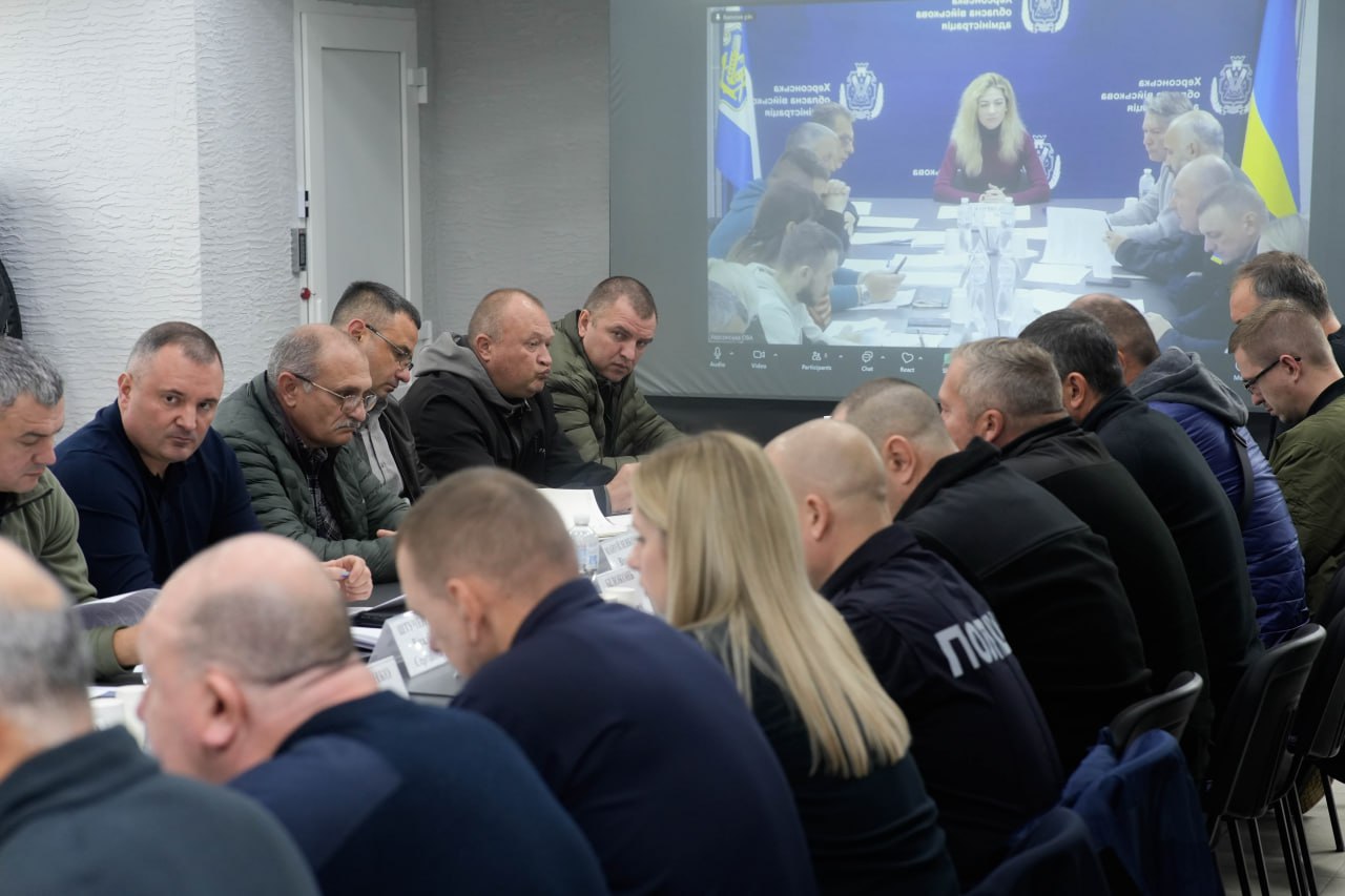 Велике зібрання людей, переважно чоловіків у темному одязі та формі (включаючи поліцейських, судячи з напису на спині одного з них), сидять за столом під час наради. На стіні позаду них демонструється зображення іншої частини цієї ж зустрічі або відеоконференції, де видно жінку зі світлим волоссям у бордовій водолазці в центрі столу на тлі логотипів Херсонської обласної військової адміністрації.
