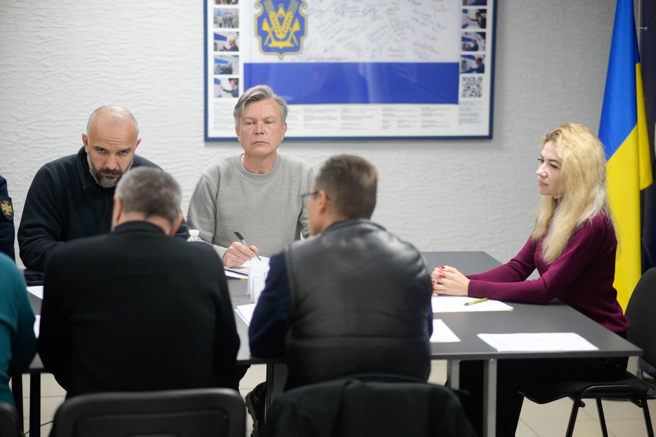 Четверо людей, включаючи чоловіка в сірому светрі та жінку зі світлим волоссям у бордовій водолазці, сидять за столом під час зустрічі. На задньому плані розташований стенд із синьо-жовтим прапором та рамка з гербом і підписами, які, ймовірно, пов'язані з Херсонською обласною військовою адміністрацією.