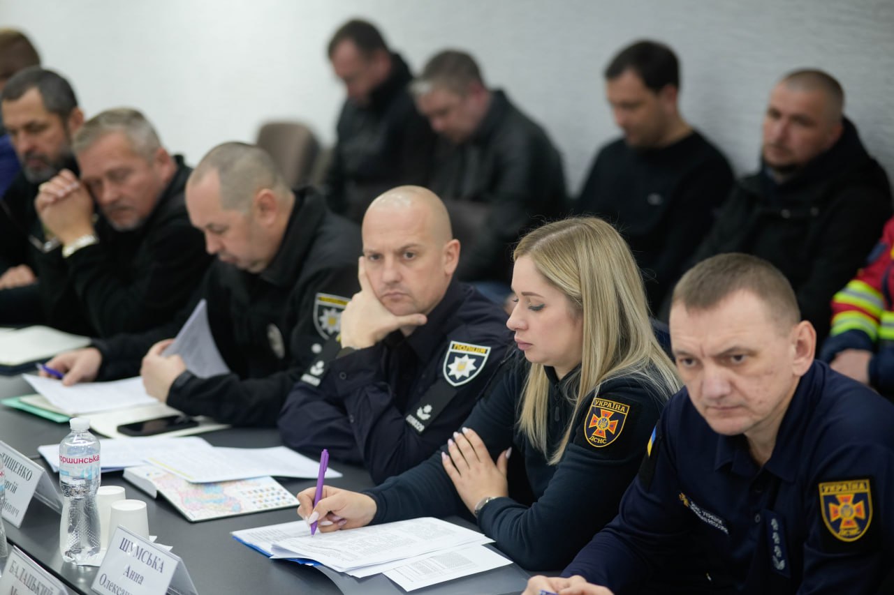 Група чоловіків і одна жінка у формі поліції та ДСНС сидять за столом під час наради. Жінка зі світлим волоссям у центрі уважно заповнює документи, поруч із нею чоловіки у формі з шевронами. На столі видно пляшки з водою, папери та іменні таблички.