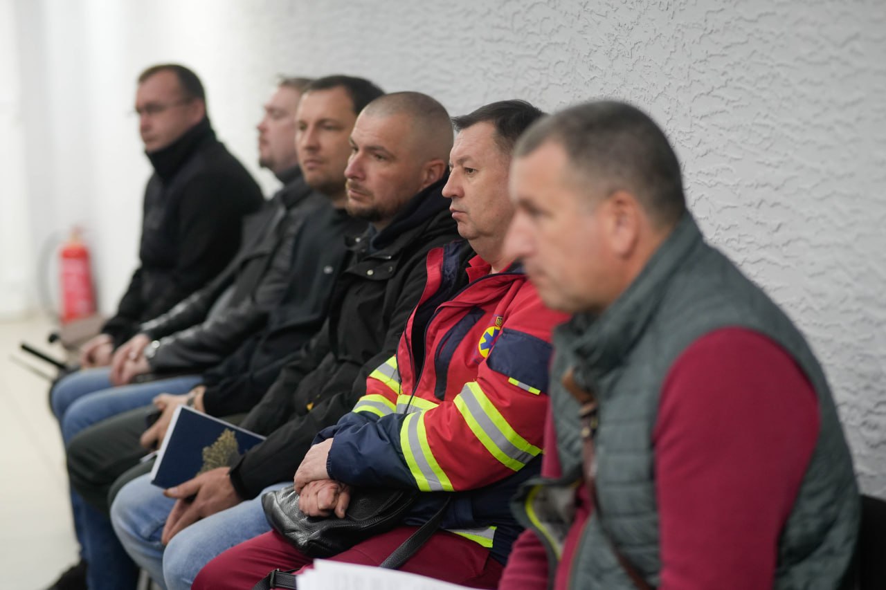 Ряд чоловіків сидить на лавці біля стіни, чекаючи. Чоловік у центрі одягнений у червоно-синю форму з жовтими світловідбивними смугами, схожу на форму працівника ДСНС або медика. Інші чоловіки одягнені в цивільний або темний одяг.