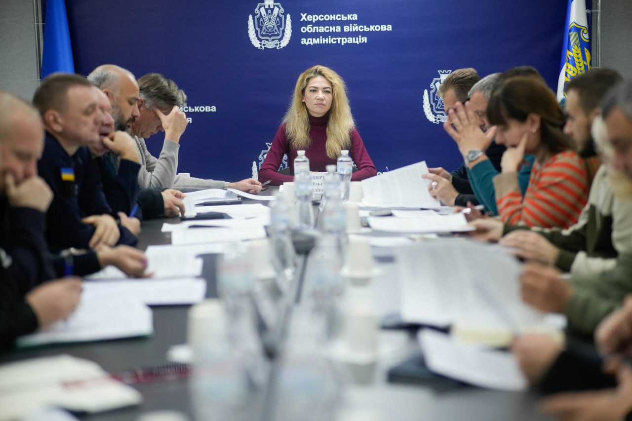 Жінка з довгим світлим волоссям у бордовій водолазці сидить у центрі великого столу, дивлячись прямо перед собою. Вона оточена багатьма людьми, які сидять навколо столу, заповненого паперами та пляшками з водою. На задньому плані розташовані прапори та логотипи Херсонської обласної військової адміністрації.