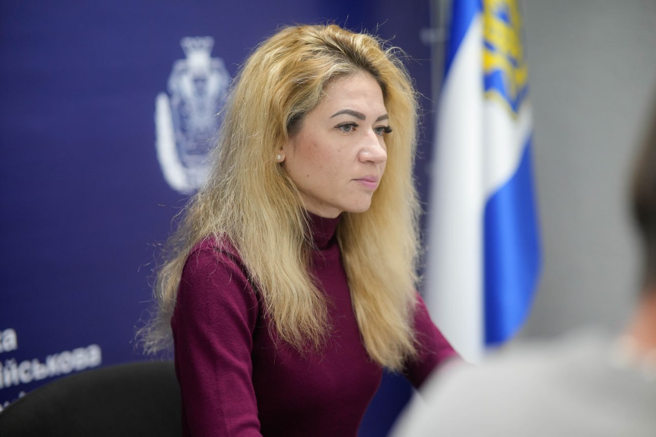 Крупний план жінки з довгим світлим волоссям у бордовій водолазці, яка серйозно дивиться праворуч від камери під час зустрічі. На задньому плані видно синю стіну з елементами логотипа Херсонської обласної військової адміністрації та частину синьо-жовтого прапора Херсонської області.