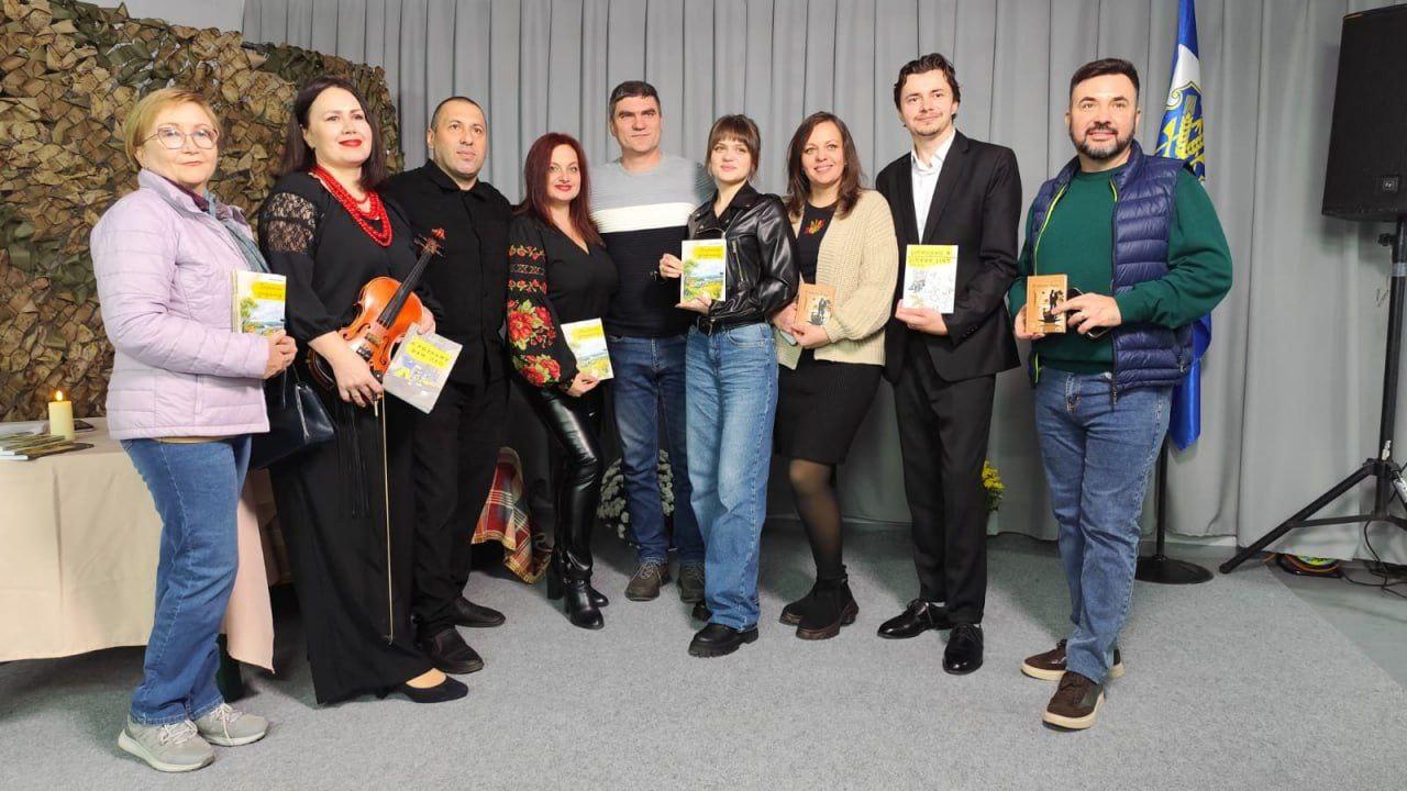 Груповий портрет восьми людей, які стоять перед сірими шторами. Більшість людей тримають у руках невеликі книжки, ймовірно, під час презентації чи літературного заходу. Серед них одна жінка тримає скрипку. На задньому плані зліва видно елемент захисної маскувальної сітки та прапор України.