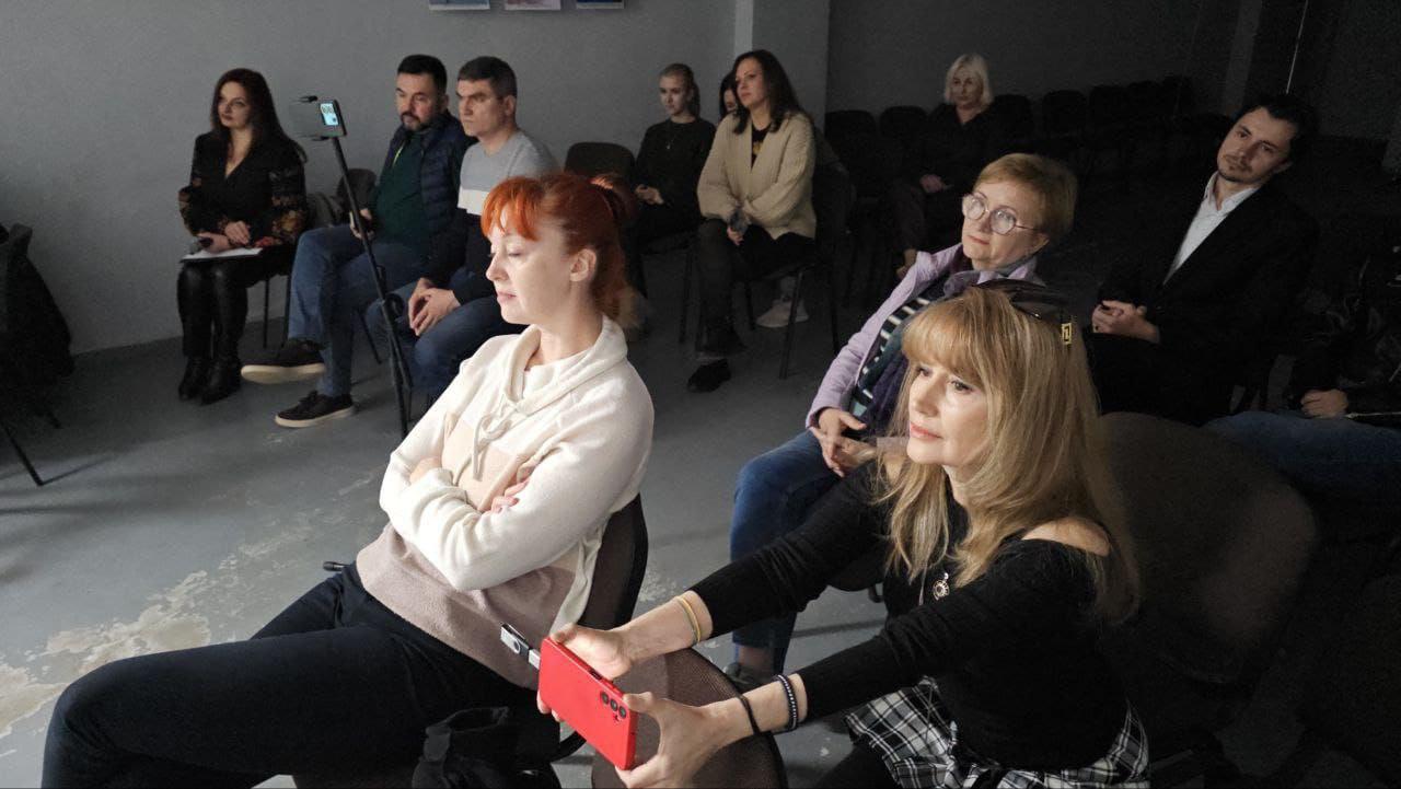 Група людей сидить у кріслах, дивлячись на сцену під час літературного заходу. На передньому плані жінка з рудим волоссям сидить зі схрещеними руками, а інша жінка з довгим світлим волоссям тримає в руках червоний телефон, роблячи знімок або відео.