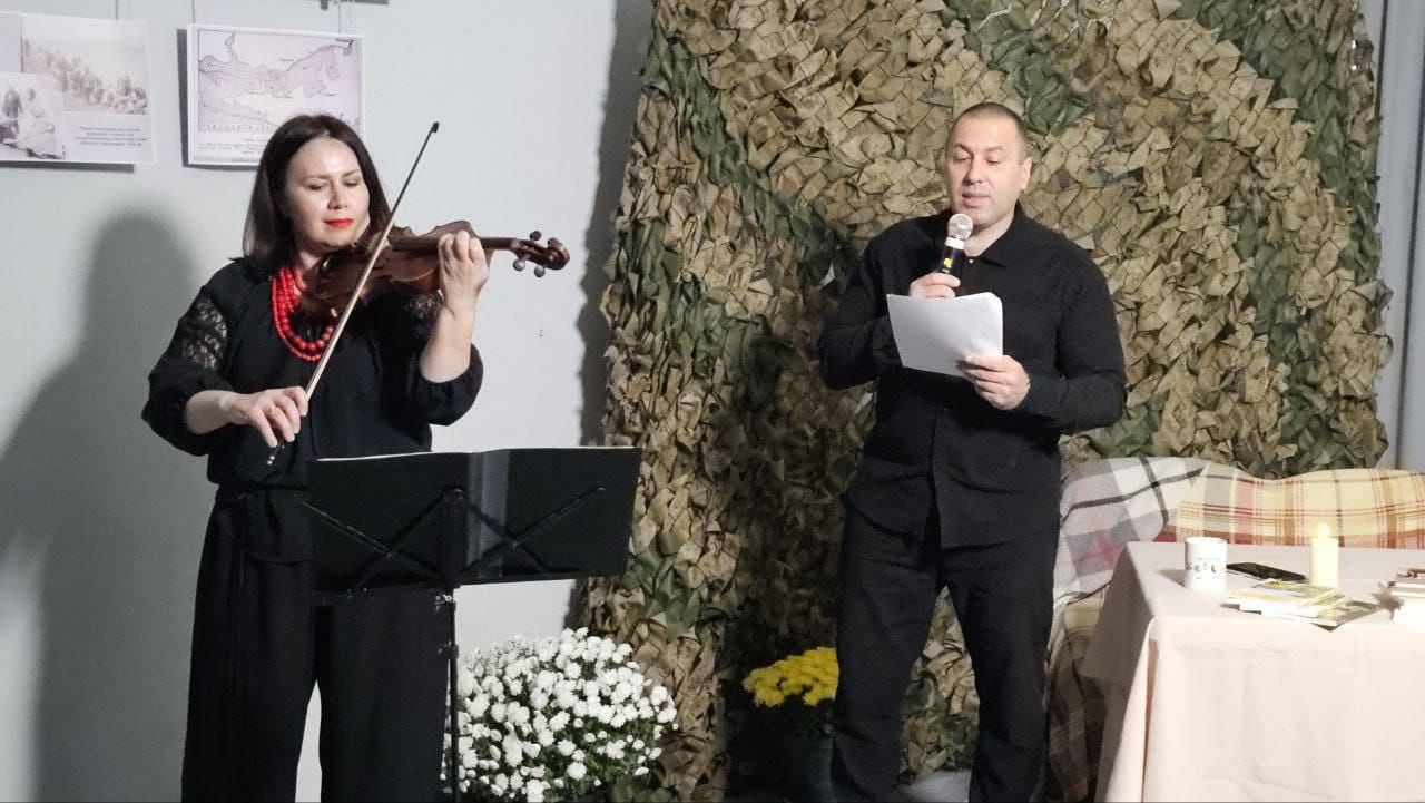 Жінка в чорному одязі та червоному намисті грає на скрипці (зліва), а чоловік у чорному одязі (справа) стоїть із мікрофоном, читаючи папір. Вони виступають на сцені перед стіною, частково покритою маскувальною сіткою, поряд зі столом із книгами та свічкою, та вазонами з хризантемами.