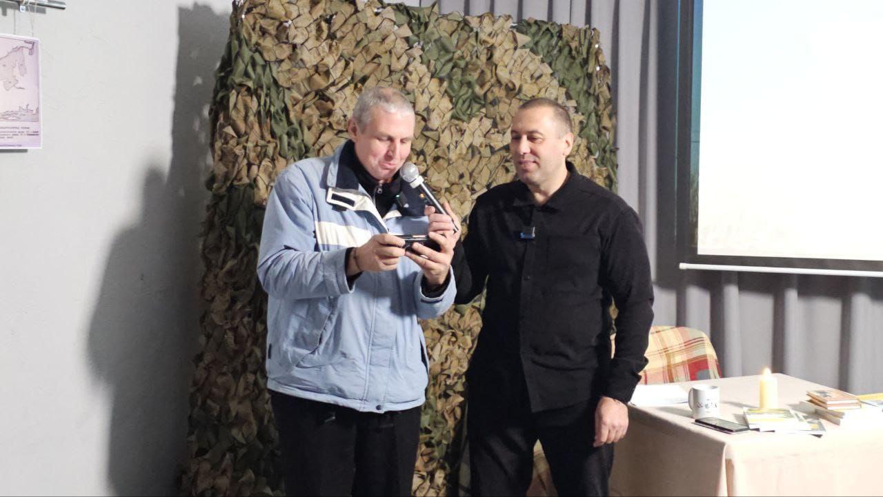 Двоє чоловіків стоять на сцені на тлі маскувальної сітки. Чоловік у світлій куртці (зліва) говорить у мікрофон, тримаючи в руках, ймовірно, невеликий телефон або записник. Чоловік у чорній сорочці (справа) стоїть поруч, дивлячись на нього.