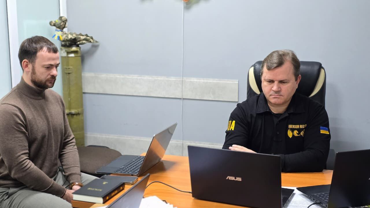 Два чоловіки сидять за столом, на якому стоять ноутбуки. Чоловік ліворуч у светрі, а праворуч — у чорній сорочці з написом KHERSON MILITARY ADM та символікою. На задньому плані видно артилерійський снаряд і прапори України.