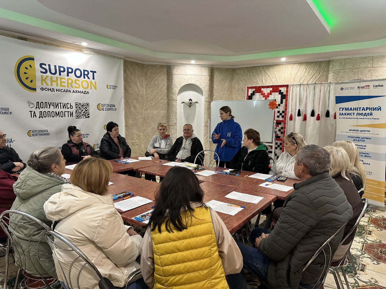 Група дорослих людей сидить за великим столом під час зустрічі чи тренінгу. Дівчина у синій кофті стоїть і виступає перед ними. На тлі видно банери SUPPORT KHERSON. ФОНД НІСАРА АХМАДА та ГУМАНІТАРНИЙ ЗАХИСТ ЛЮДЕЙ (Caritas).